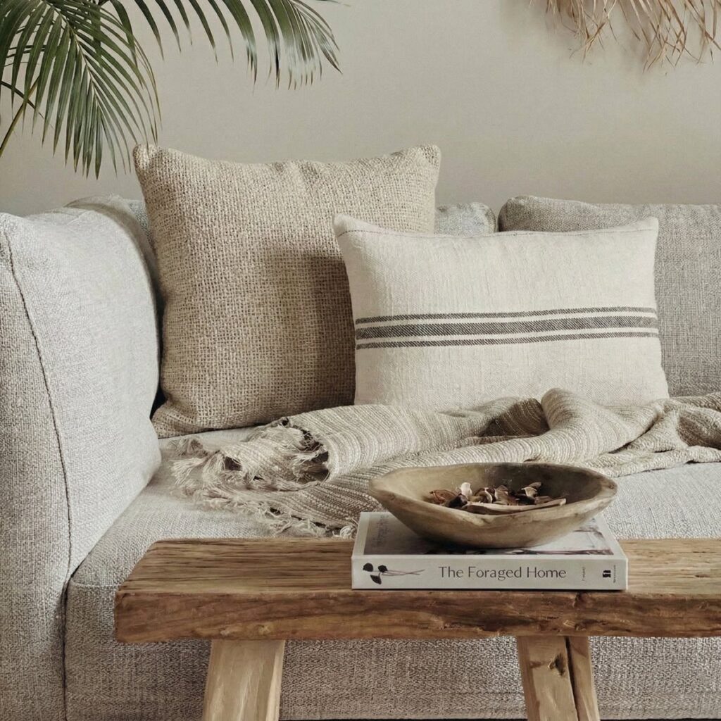 Close-up of a light grey sofa styled with beige burlap and striped linen pillows. In the foreground, a raw wood bench features a wooden bowl with dried botanicals and the book 'The Foraged Home'. A green palm leaf frames the left side.