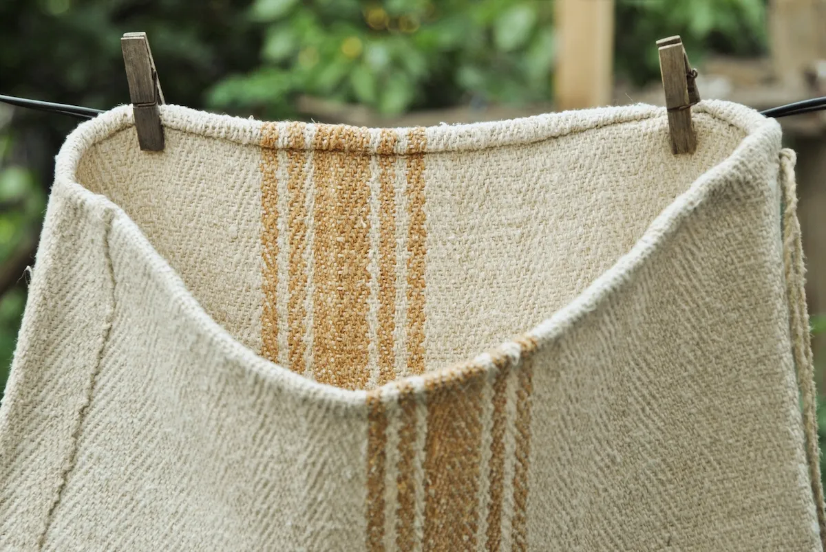 Antique grain sack hanging on clothesline outdoors, unbleached natural hemp in warm yellow-oatmeal tone, caramel woven stripes, 4-shaft twill weave, Transylvania