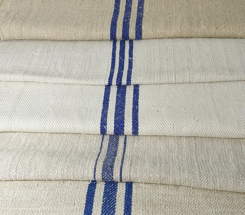Antique grain sack fabric showing herringbone and diagonal twill weave structure, oatmeal handspun hemp and cotton with blue woven stripes, dense hand-woven texture visible, Transylvania circa 1900