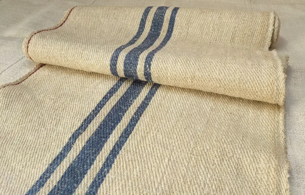 Stack of rolled antique grain sack fabric pieces sourced from Transylvania, oatmeal handspun hemp with navy blue woven stripes, herringbone twill weave, awaiting restoration