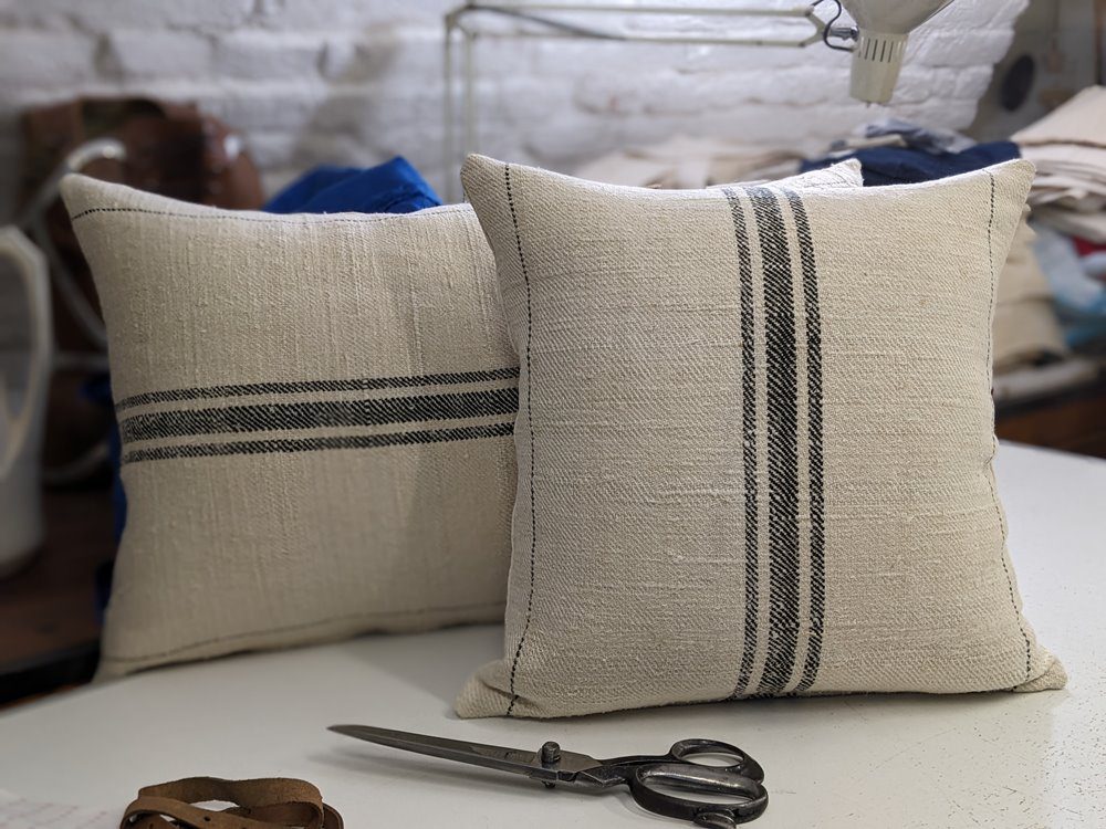 Two handmade decorative pillows crafted from vintage Transylvanian grain sacks featuring signature black stripes, displayed in a studio setting with vintage scissors.