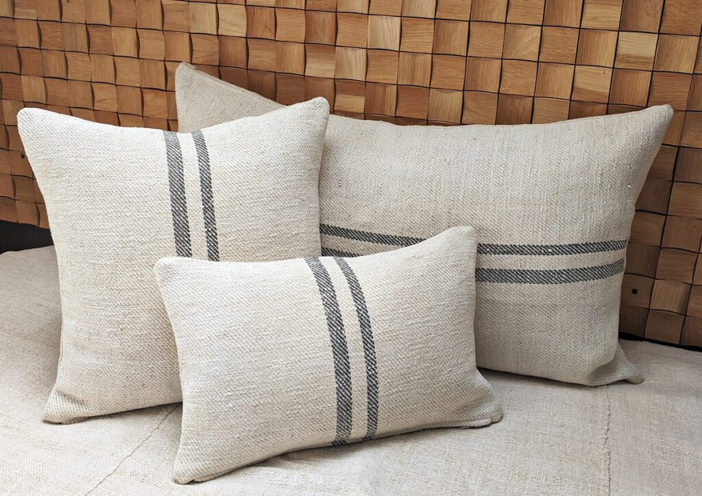 A set of three vintage-style decorative cushions in various sizes, featuring black stripes on natural hemp or linen fabric, arranged on a textured sofa.