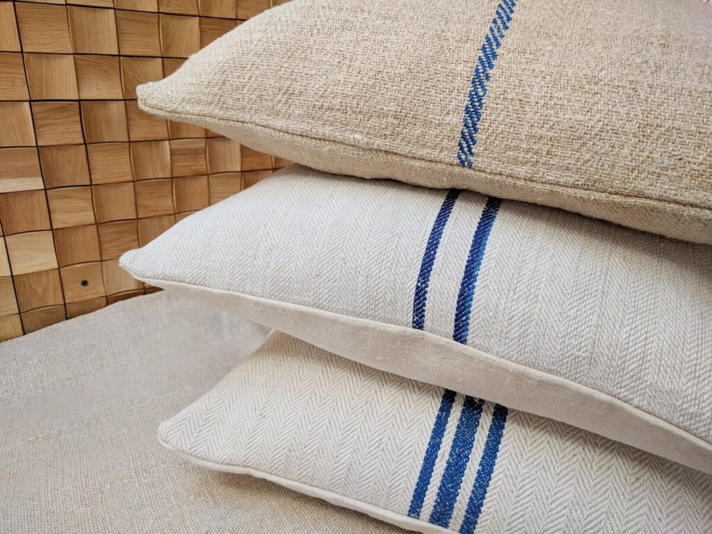 Close-up detail of three stacked decorative pillows made of rustic beige linen with vertical blue stripes against a wooden panel background.