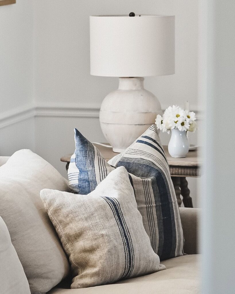 antique grain sack pillows with blue stripes on linen sofa, ceramic lamp and white daisies, editorial interior styling