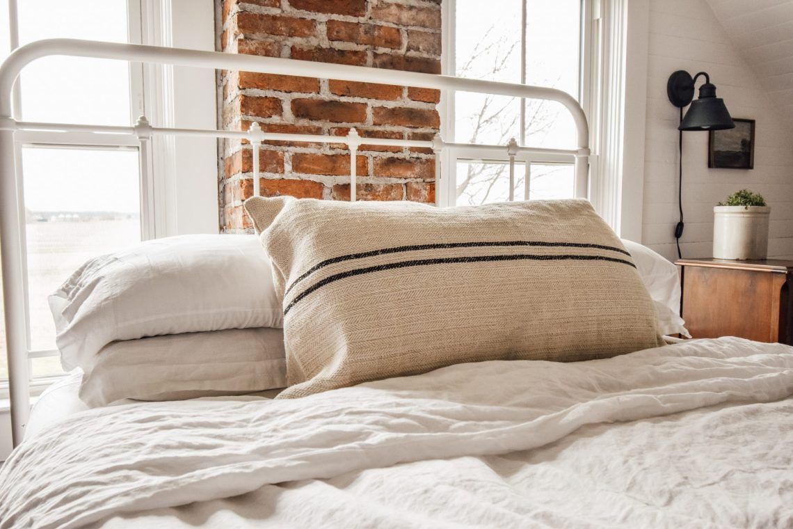 Antique grain sack lumbar pillow on white iron bed, exposed brick wall, natural light, black stripe hemp textile, bedroom styling